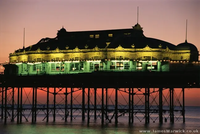 Historic pier pavilion illuminated in gold and green lights at dusk, reflected in shallow water below.