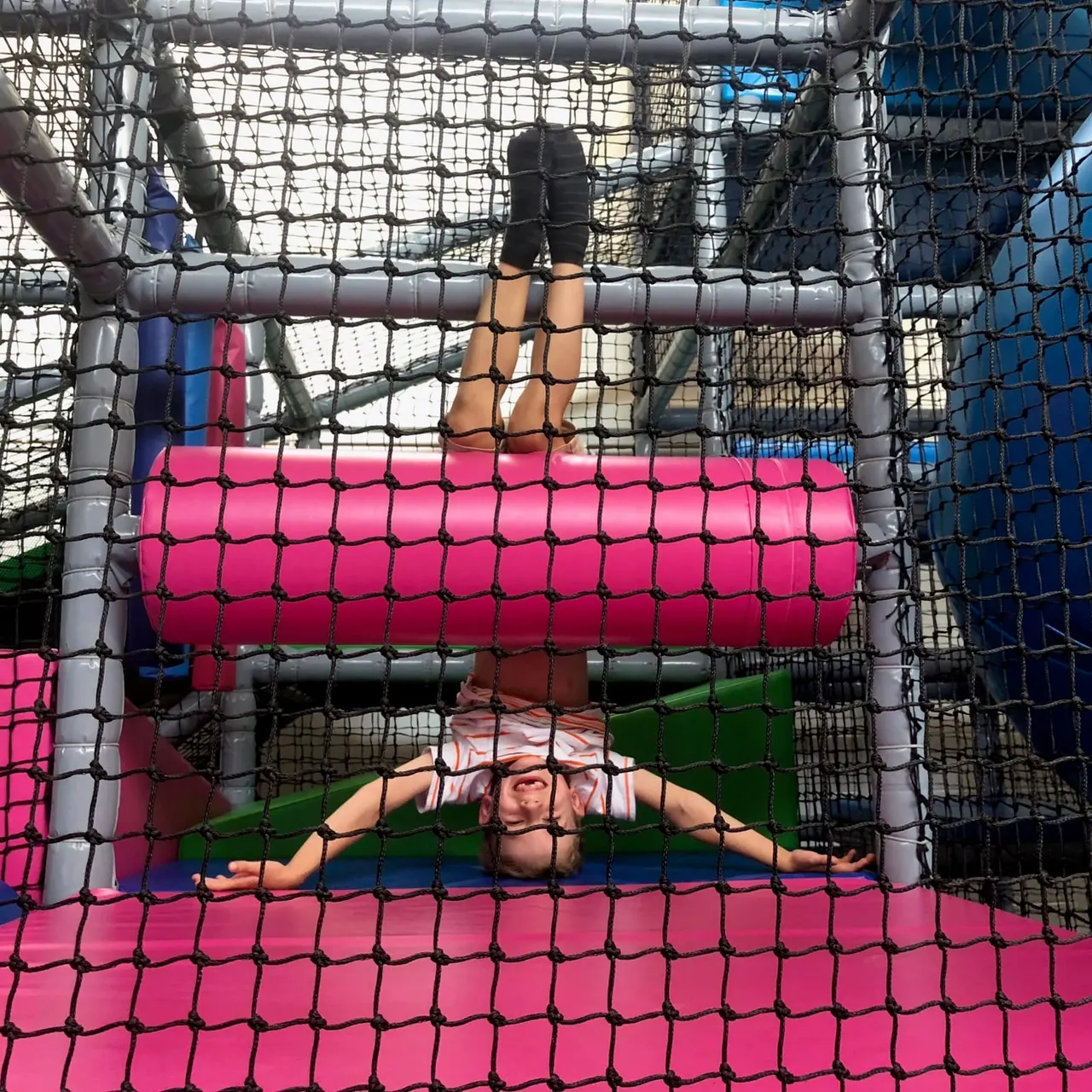 Child playing on a bright pink inflatable roller inside a netted soft play structure.