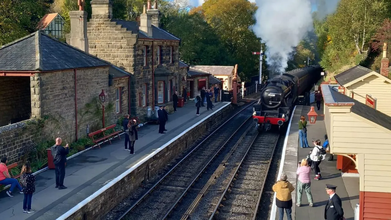 Steam locomotive pulling carriages into a heritage railway station with visitors and period buildings.