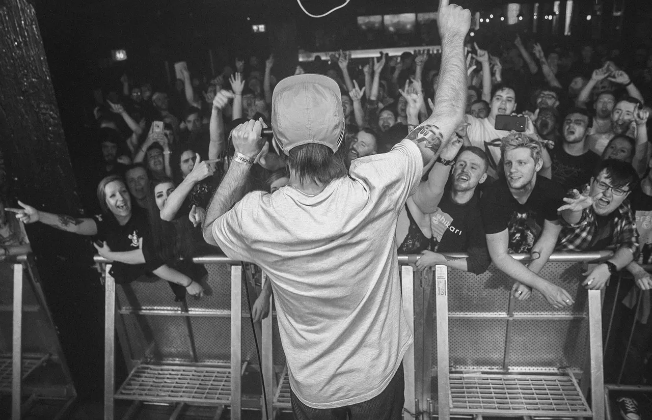 Performer on stage with arms raised facing a large crowd of enthusiastic spectators behind a barrier at a nightclub venue.