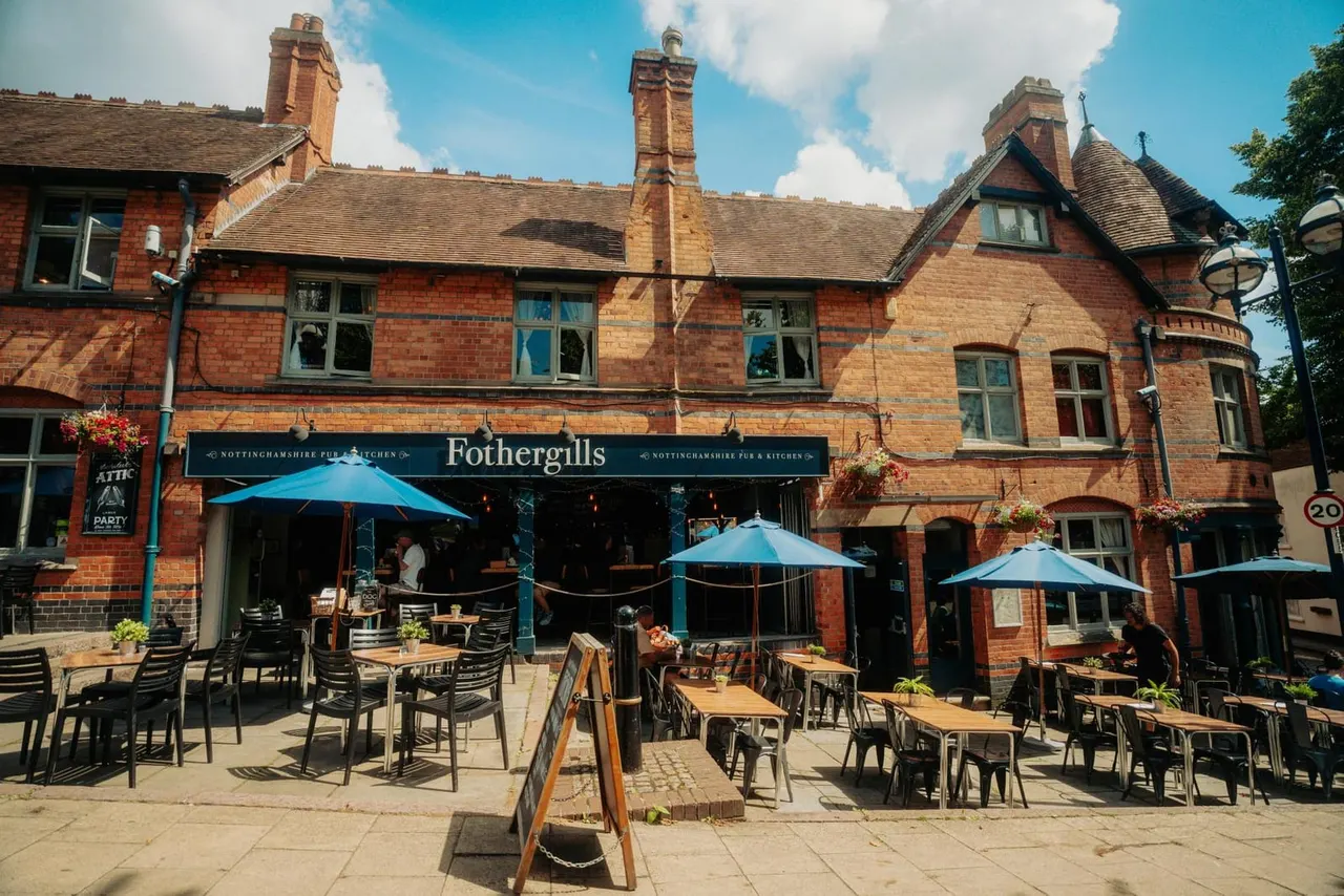 A red-brick Victorian pub with blue umbrellas and wooden tables on an outdoor patio on a sunny day.
