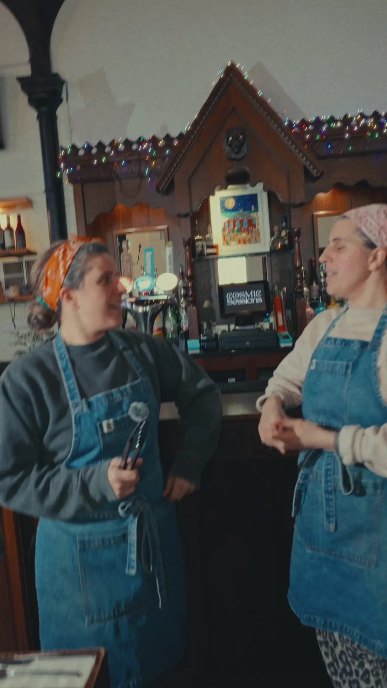 Two staff members in denim aprons converse inside a festively decorated gingerbread house-themed kitchen venue with colourful garland and Christmas decorations.