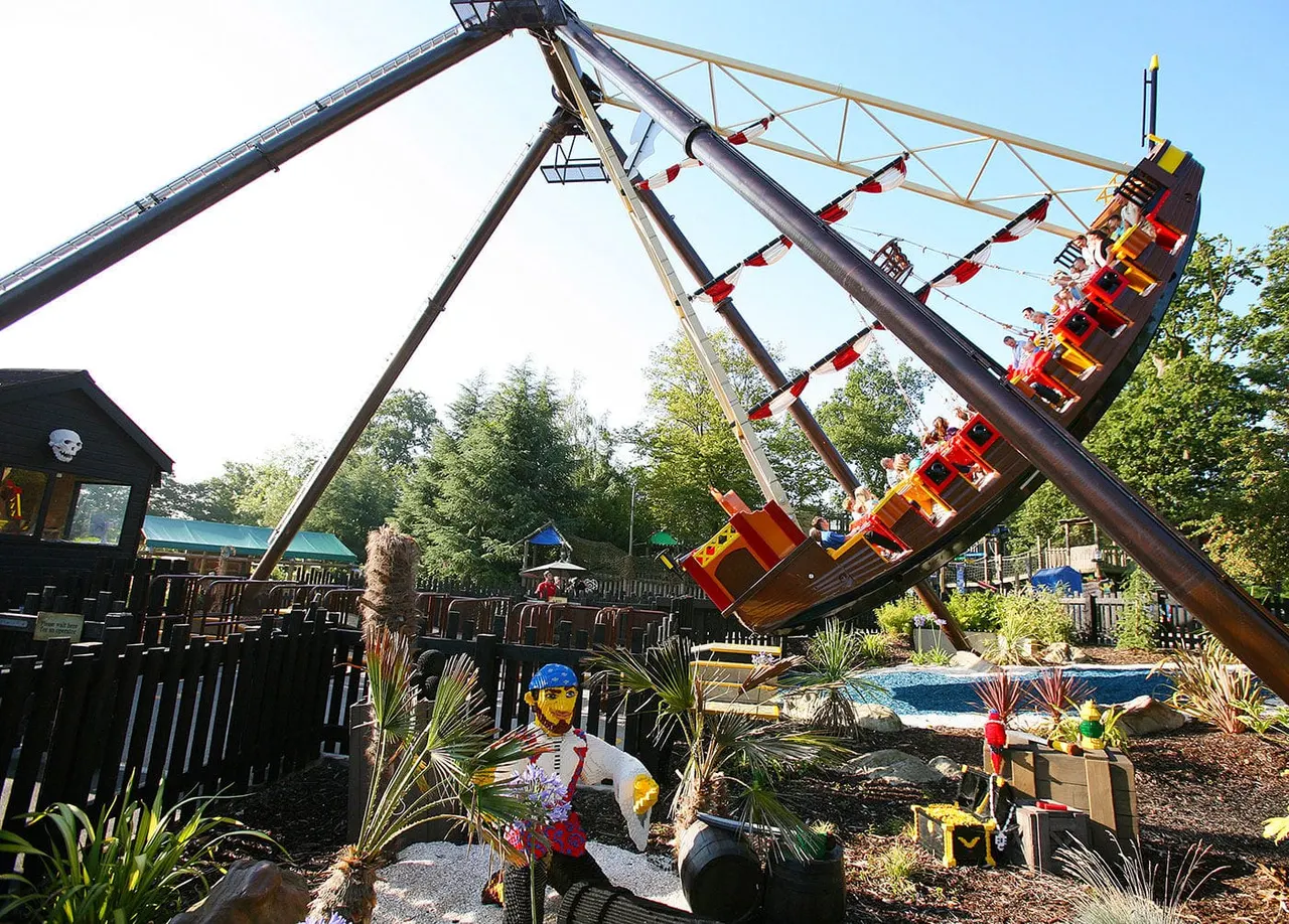 Red and yellow pirate ship ride swinging high with passengers, surrounded by themed playground structures and tropical plants.