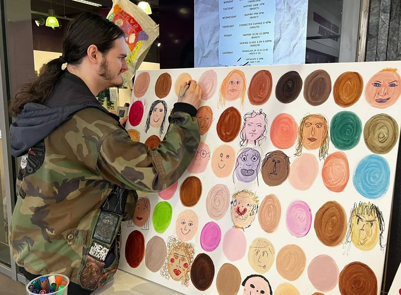 Man in camouflage jacket painting a face on a large collaborative artwork featuring numerous hand-drawn faces and coloured circles.