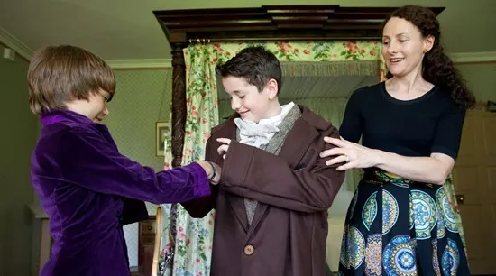 A woman helps two young children try on Victorian-era clothing indoors.