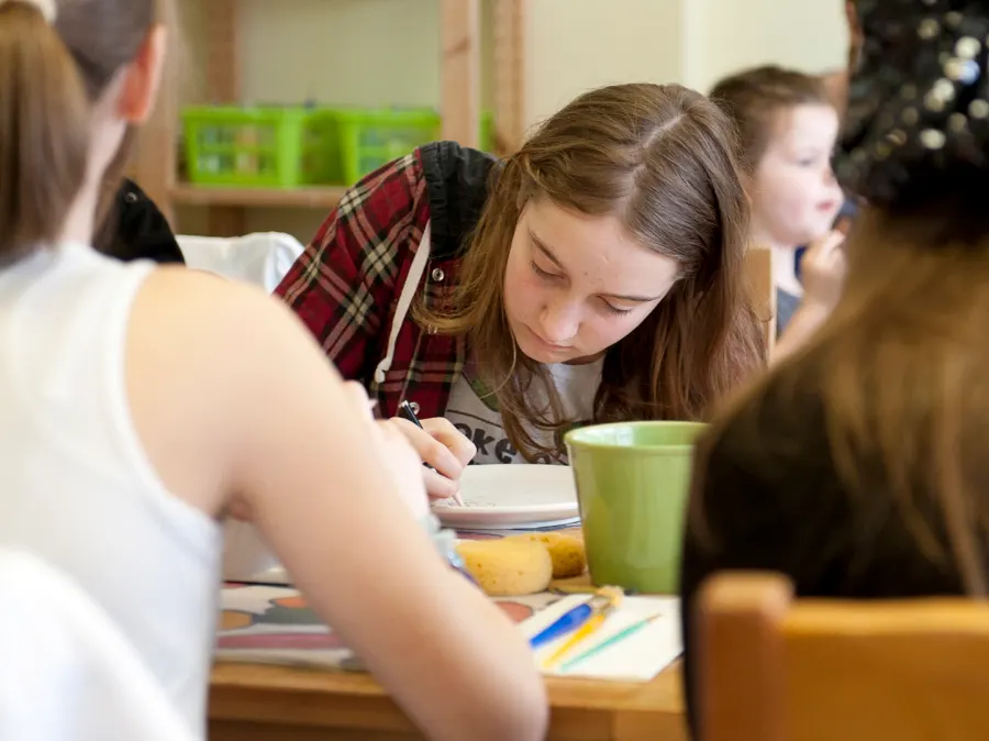 This is a PHOTOGRAPH, not a designed graphic.

Children seated at a table during an activity, with one girl in a tartan shirt concentrating on her work, surrounded by craft supplies and a green cup.