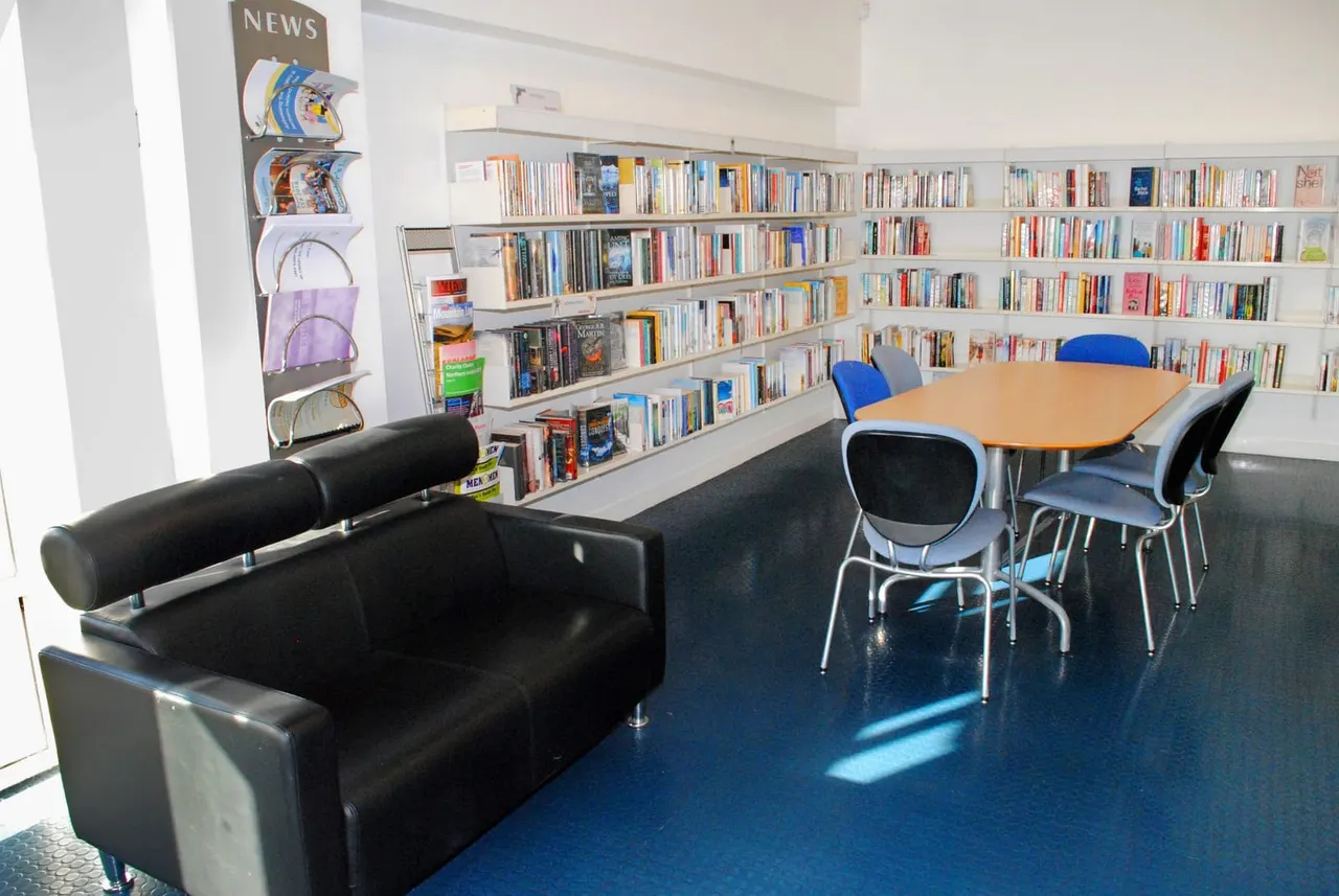 Library reading room with bookshelves, a black sofa, wooden table with blue chairs, and storage baskets on wall shelving.