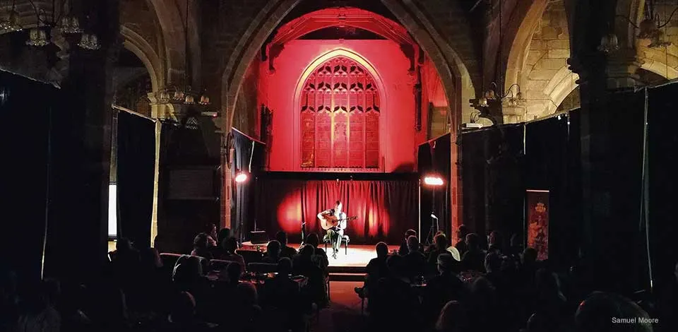 A musician performs on stage in a historic stone church with red lighting, whilst an audience watches from seated rows.