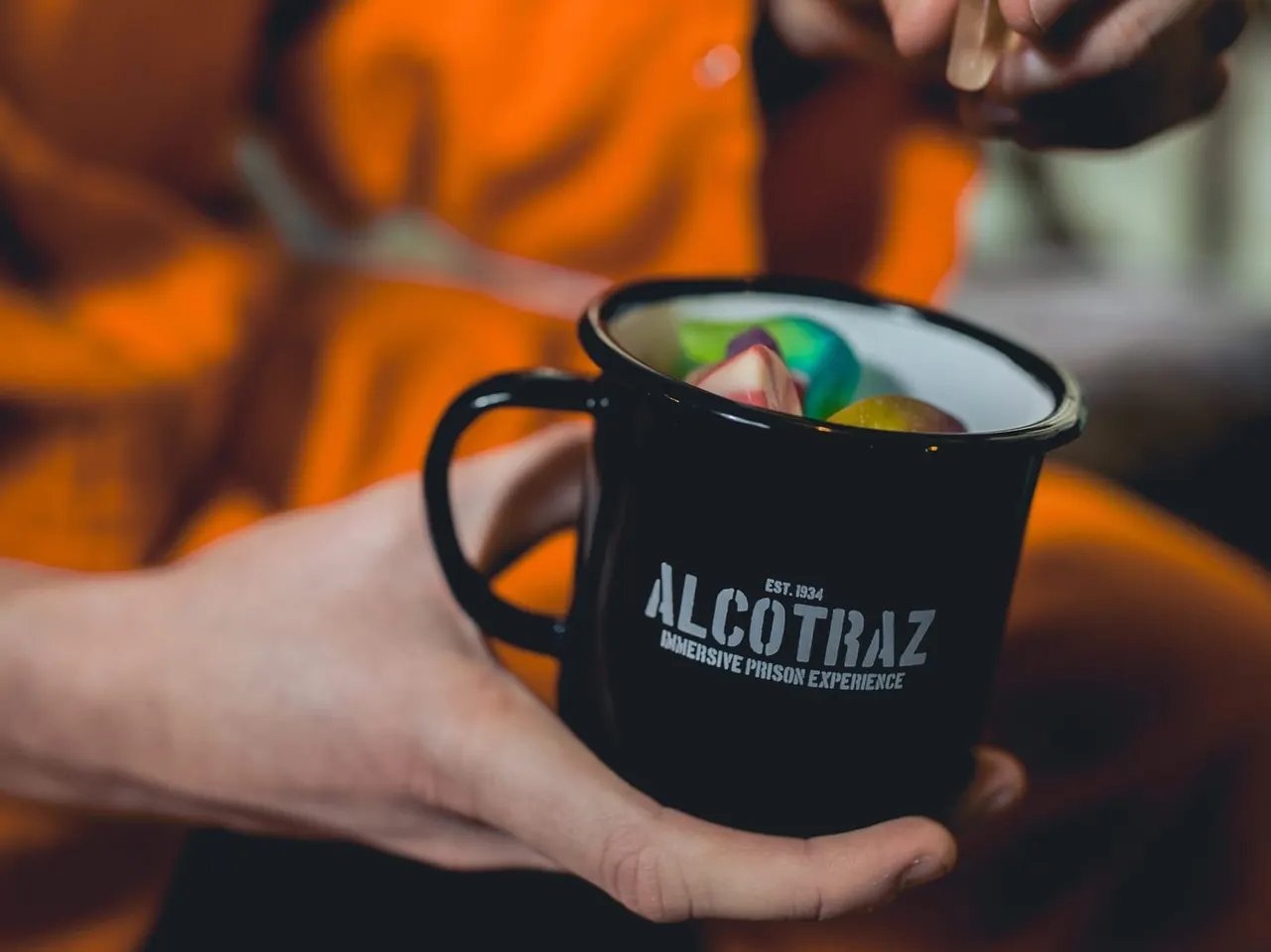 Person holding a black enamel mug containing a colourful cocktail at an indoor venue.