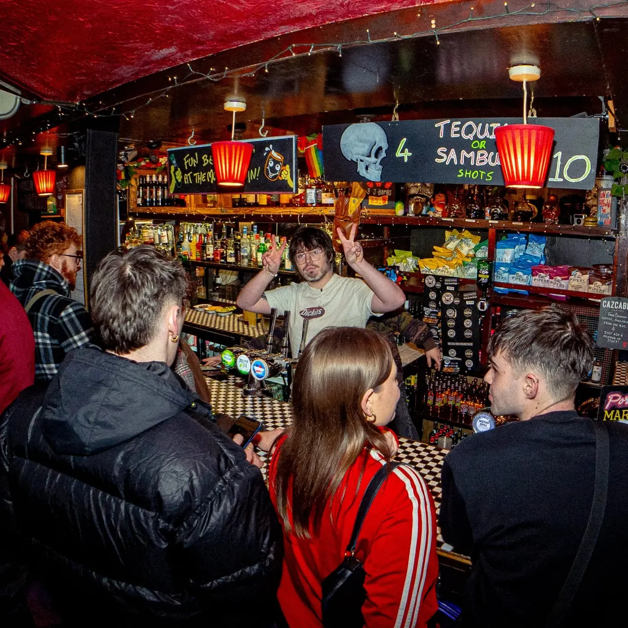 Bartender making drinks for a group of customers at a crowded, vibrant bar with red lanterns and skull decorations.