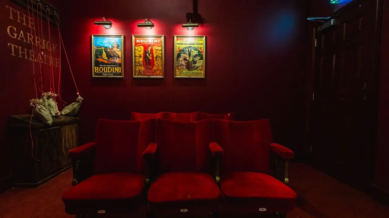 Theatre-style seating area with red velvet chairs facing a deep red wall decorated with vintage Houdini posters and suspended puppets.