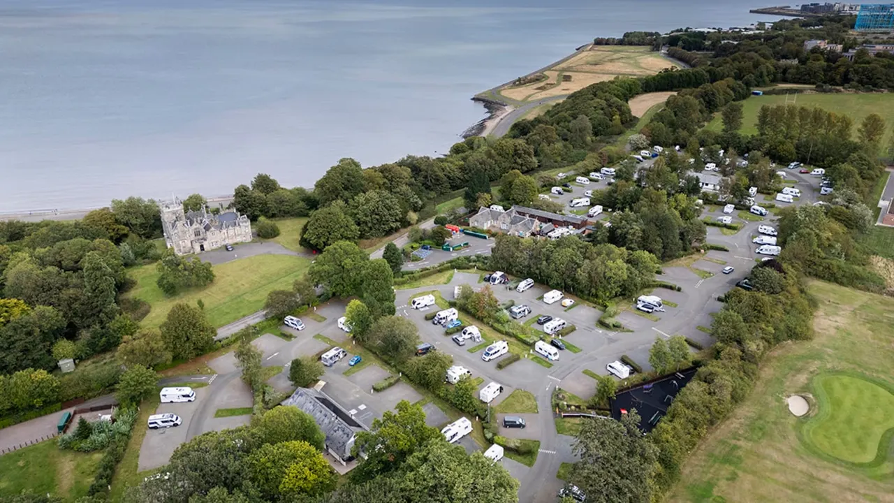 Aerial view of a caravan and motorhome campsite set in wooded parkland beside a castle and coastal estuary.
