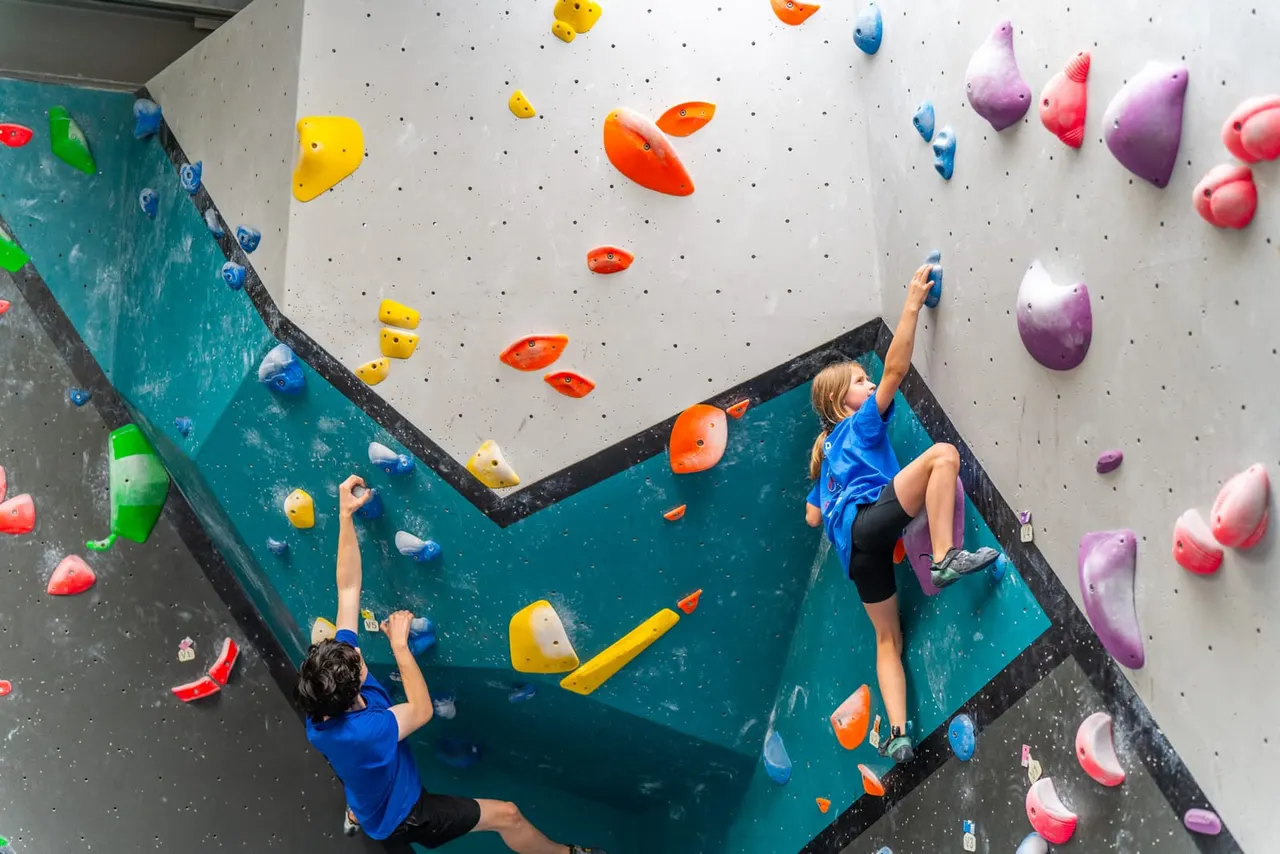 Two children climbing an indoor rock climbing wall with colourful handholds, viewed from above.