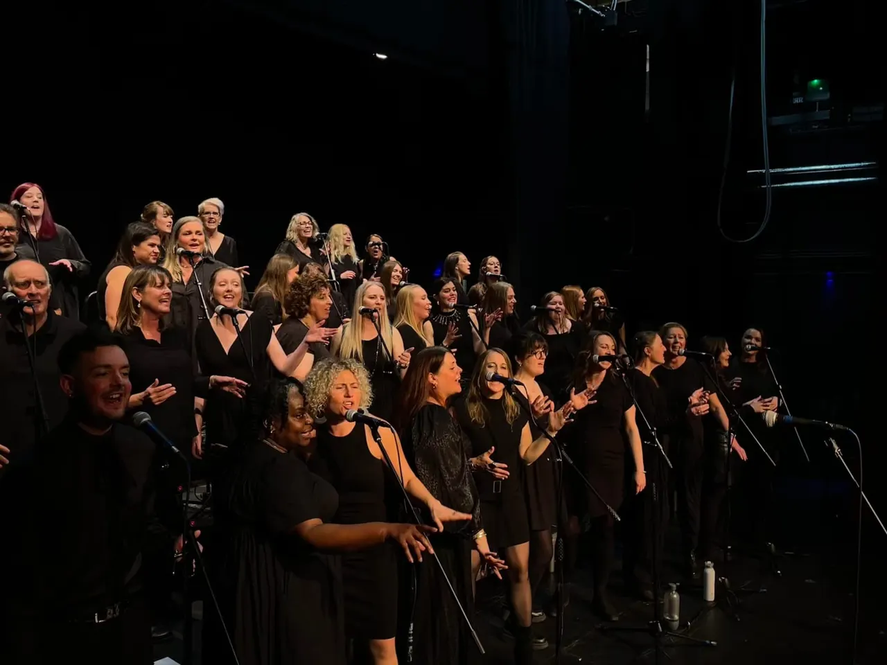 Large choir of people in black clothing performing together on an illuminated stage with microphones.