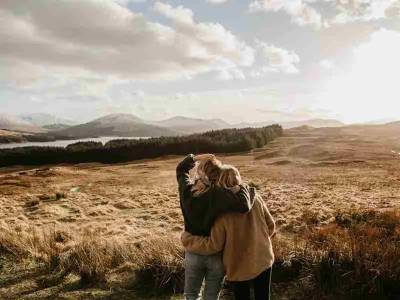 Two people embracing on moorland overlooking mountains, forests and a loch under a cloudy sky.