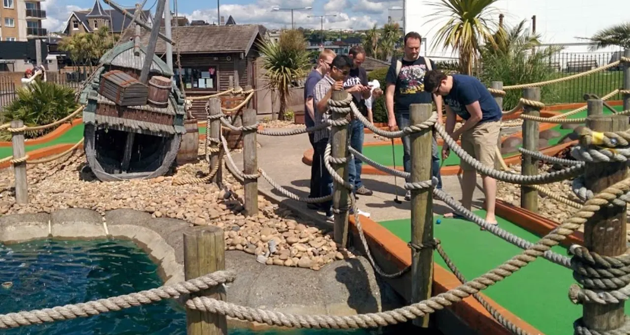 Family and adults playing mini golf on a pirate-themed course with rope barriers and themed structures.