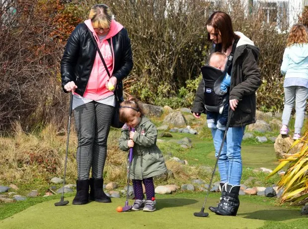 Adults and children playing mini golf on a putting green outdoors.