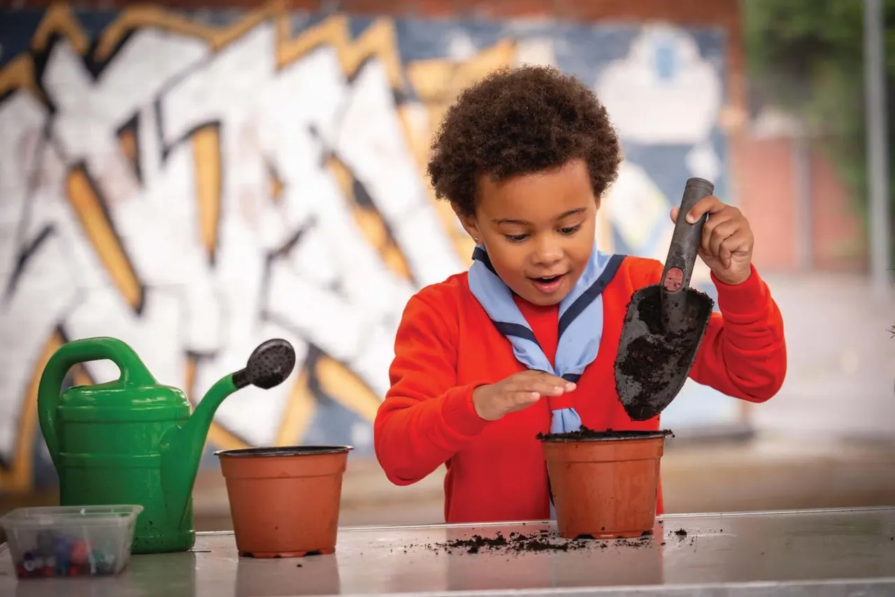 A young child in a red jacket plants seeds in a terracotta pot with a small shovel during a gardening activity.
