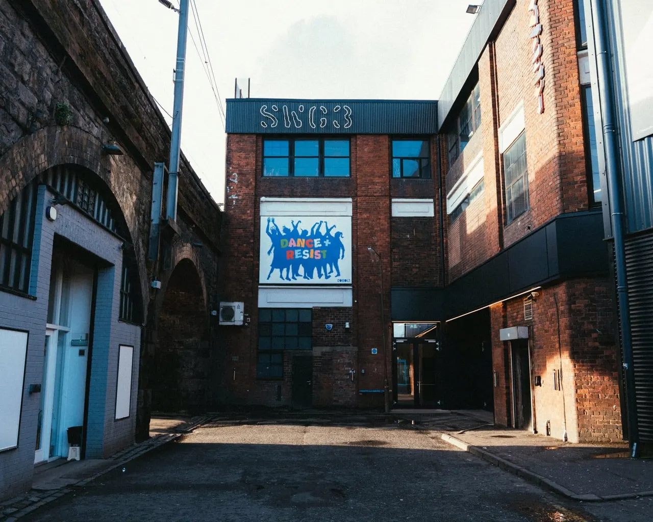 Brick industrial building with blue signage displaying "Dance + Resist" mural in a urban courtyard setting.