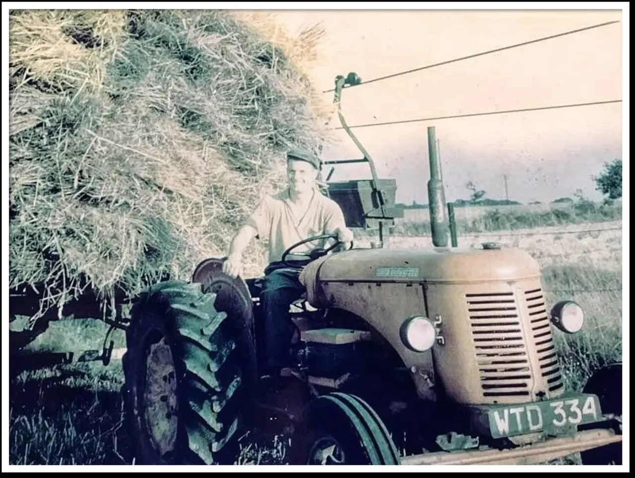 Man operating a vintage tractor loading hay onto a farm vehicle in a rural field.