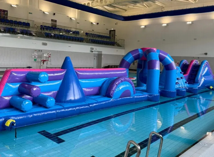 Inflatable obstacle course in bright blue and pink set up in an indoor swimming pool with spectator seating visible above.