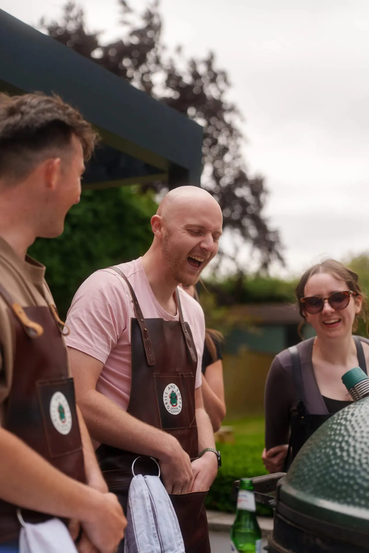 Staff members in branded aprons laugh together during an outdoor cooking class activity.