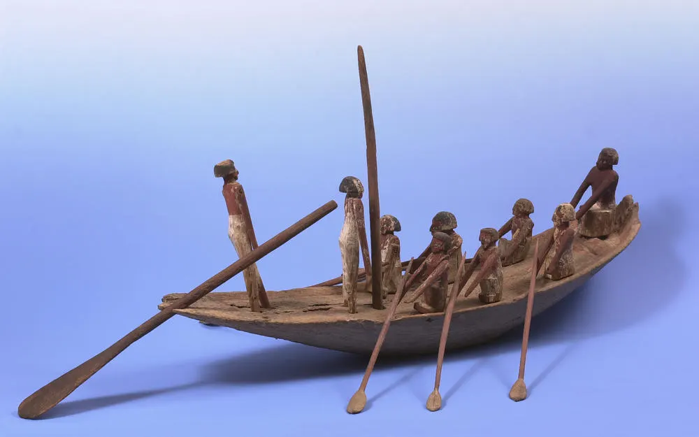 Wooden carved model of an ancient boat with multiple human figures and oars against a blue background.