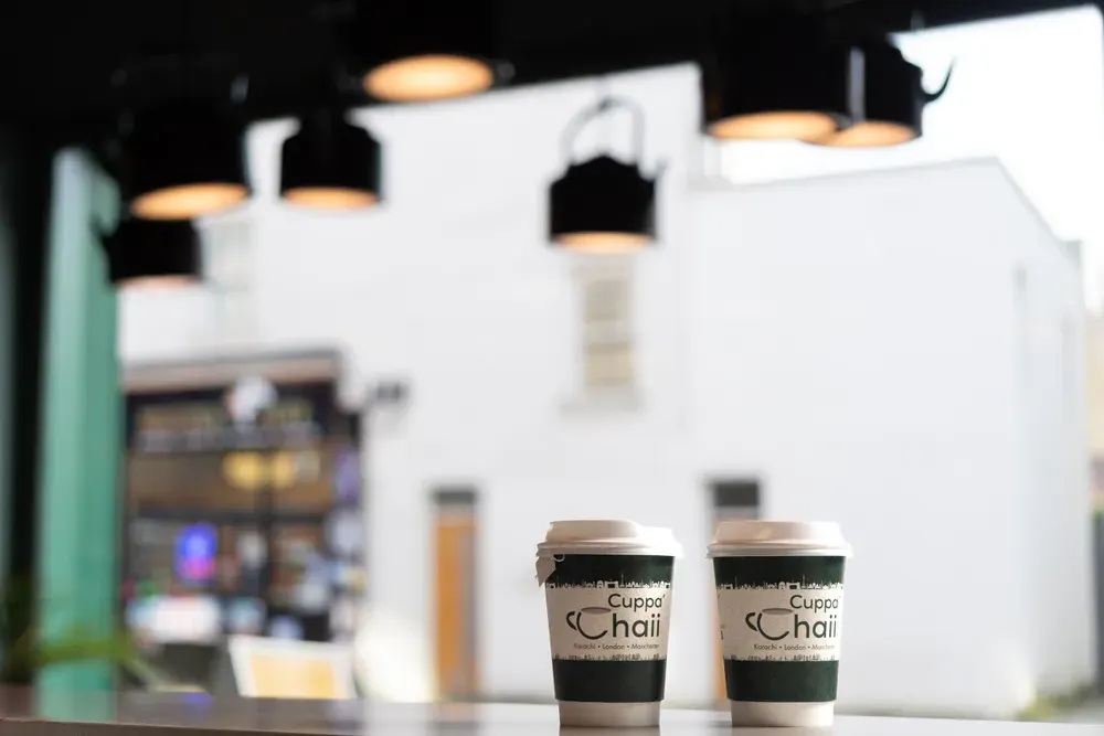 Two takeaway cups of Cuppa' Chaii chai on a counter with the café storefront blurred in the background.