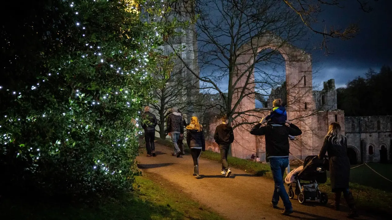 Families and visitors walking at dusk past illuminated trees and historic ruined abbey buildings.