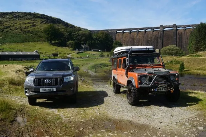 Two four-wheel-drive vehicles parked beside a river with a viaduct and green hillside behind them.