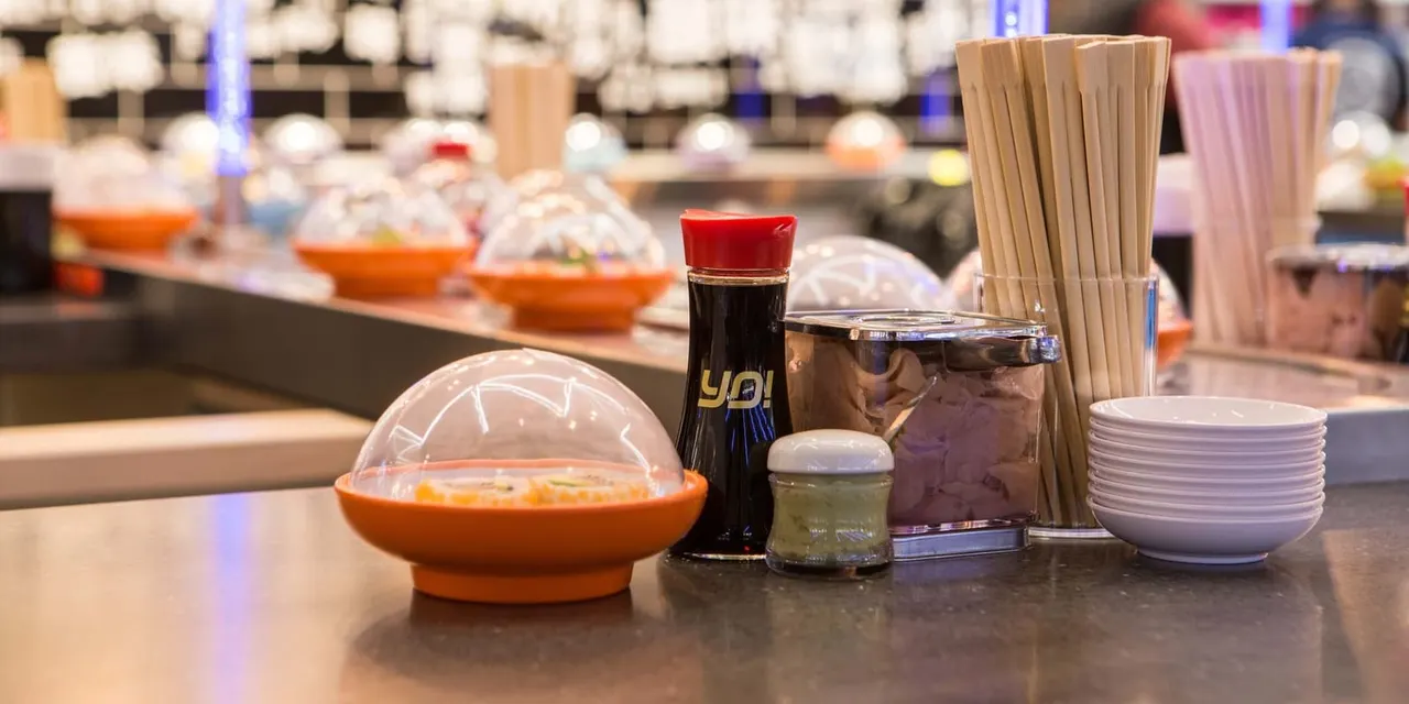 Sushi counter setup with condiments, sauce bottle, straws, bowls and orange serving dishes with dome covers.