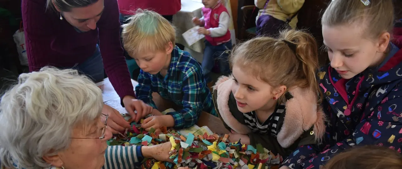 Children and an elderly woman gather around a colourful craft activity involving coloured paper strips at an indoor community event.