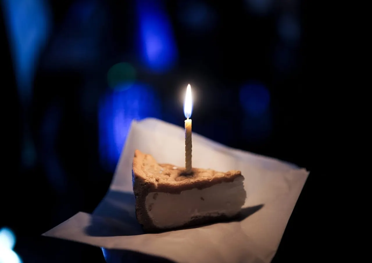 A slice of cake with a lit candle on a white napkin against a dark blue-lit background.