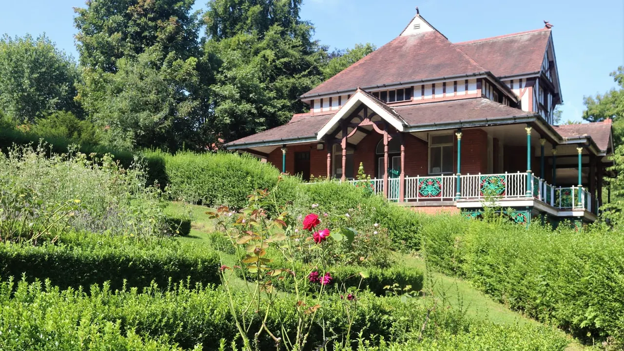 Historic red-brick building with turrets set amongst lush green gardens with flowering shrubs and mature trees.