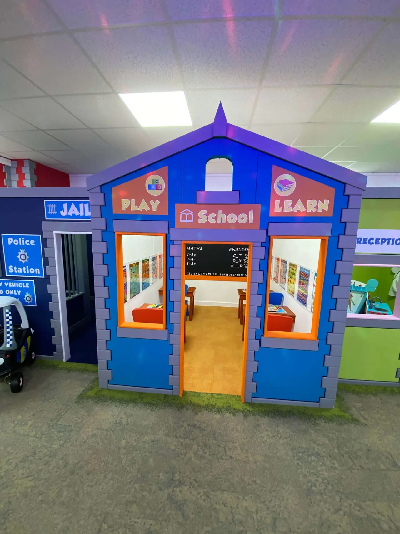 A large blue and orange play school building with windows showing educational activities inside, alongside a police station playhouse in an indoor play area.