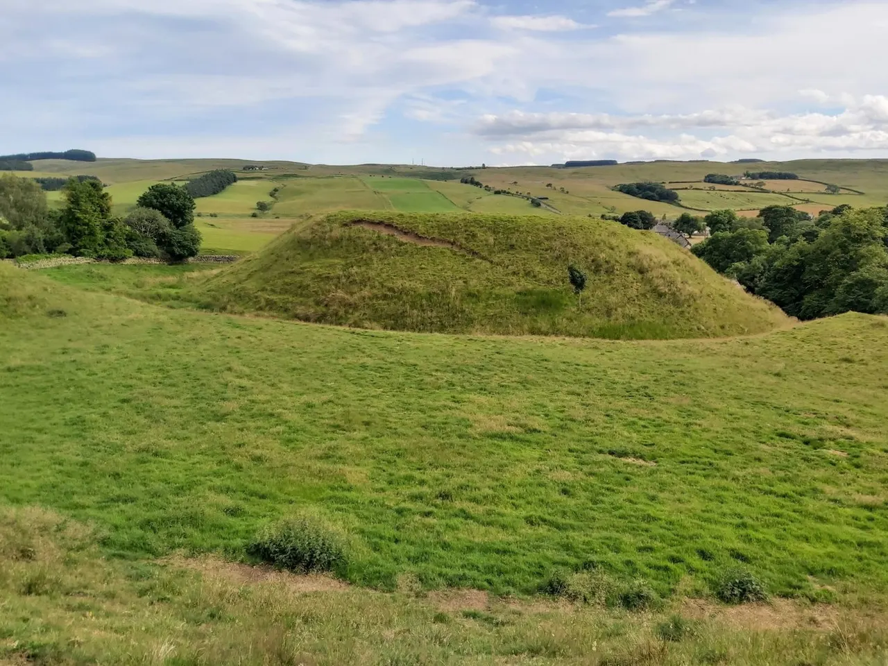 A large grass-covered earthwork mound dominates a rolling green hillside landscape beneath a cloudy sky.