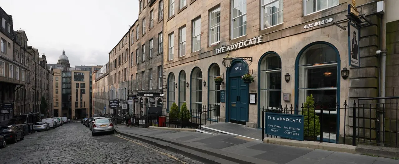 Historic Edinburgh street with stone buildings, the Advocate pub with blue door, and parked cars.