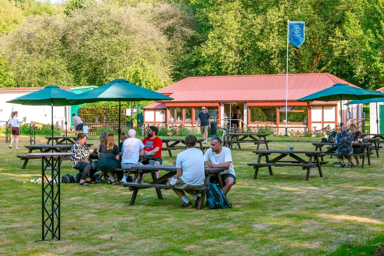 Visitors seated at picnic tables on a grass area with blue umbrellas, a clubhouse building and tree-lined background.