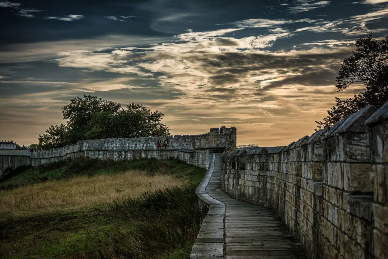 Wooden boardwalk between stone fortress walls at a historic fortification with dramatic cloudy sky above.