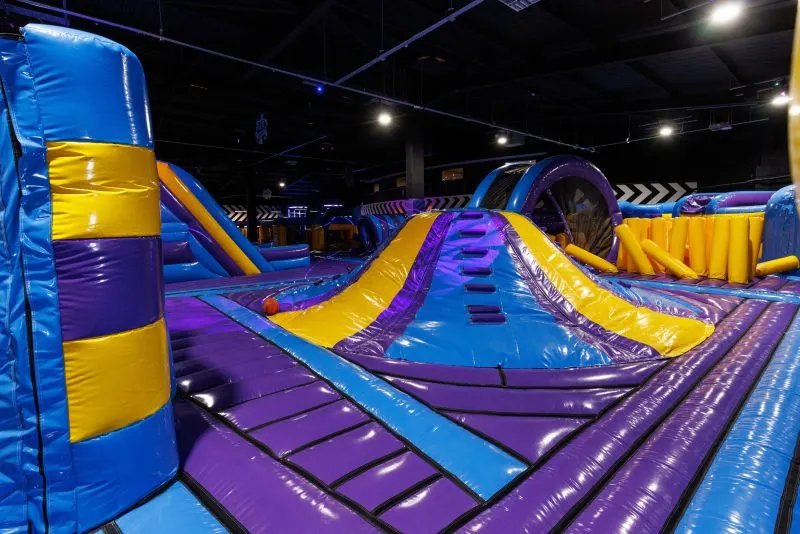 Colourful inflatable obstacle course and slides in a large indoor play arena with blue, yellow and purple padding.