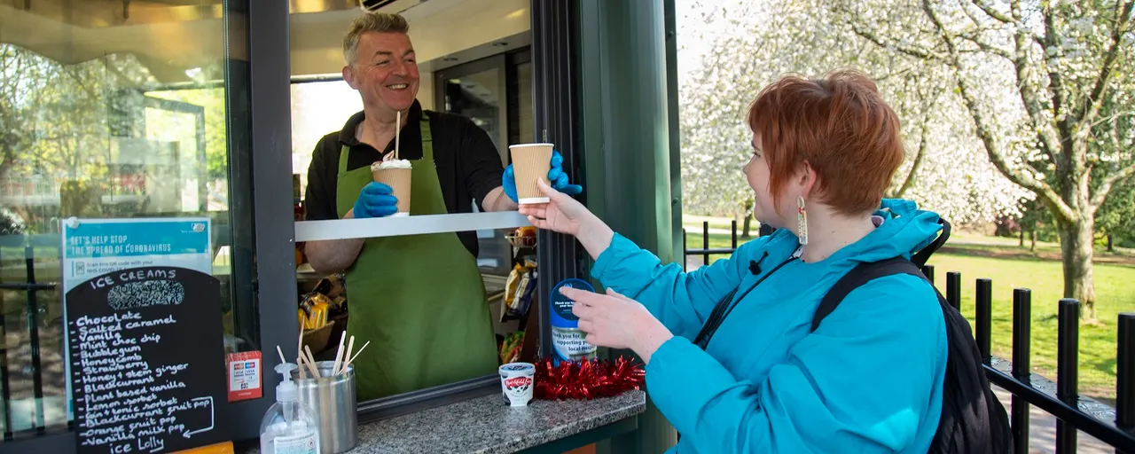 Server in green apron handing ice cream to customer at outdoor window counter, with blossoming trees visible in background.