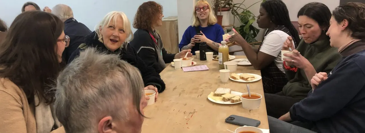 A diverse group of people of various ages gathered around a wooden table sharing food and drinks in an indoor social setting.