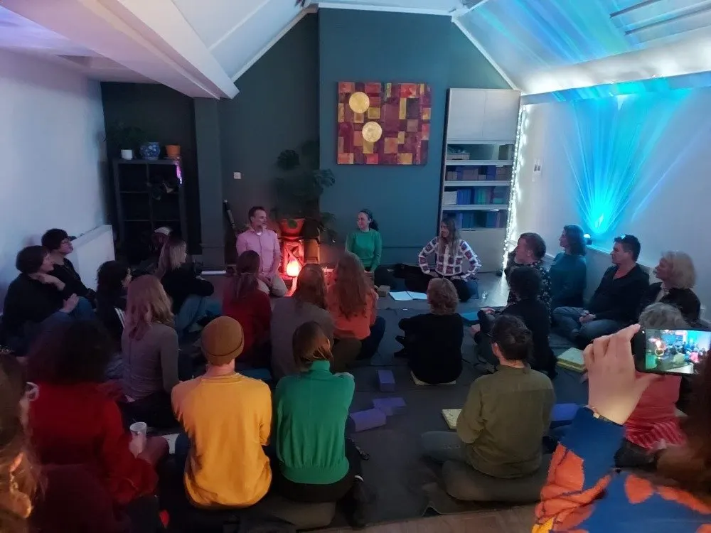 A group of adults and children sit in a circle on the floor during an indoor event with ambient blue lighting and a glowing fire feature.