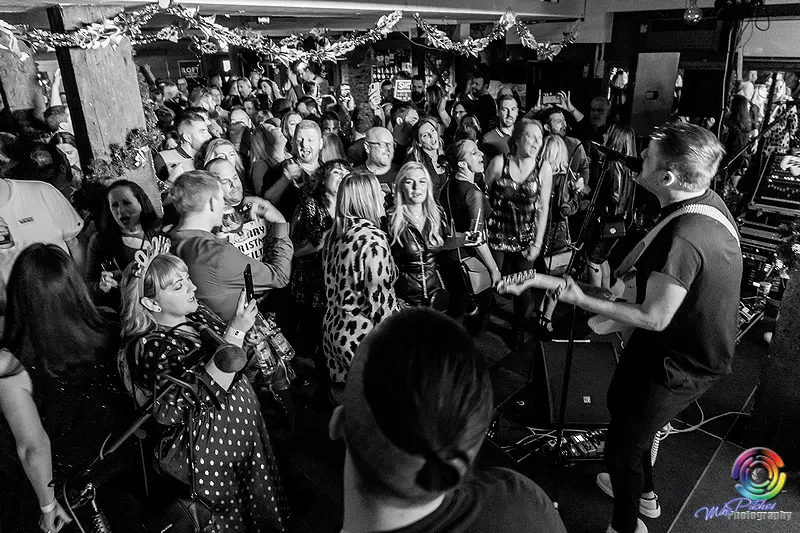 Crowded indoor bar at night with people dancing and socialising around a performer on stage, festive decorations overhead.