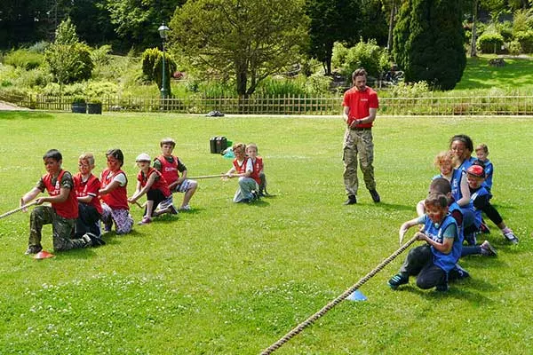 Children in red and blue teams play tug-of-war on a grass field during an outdoor activity session.