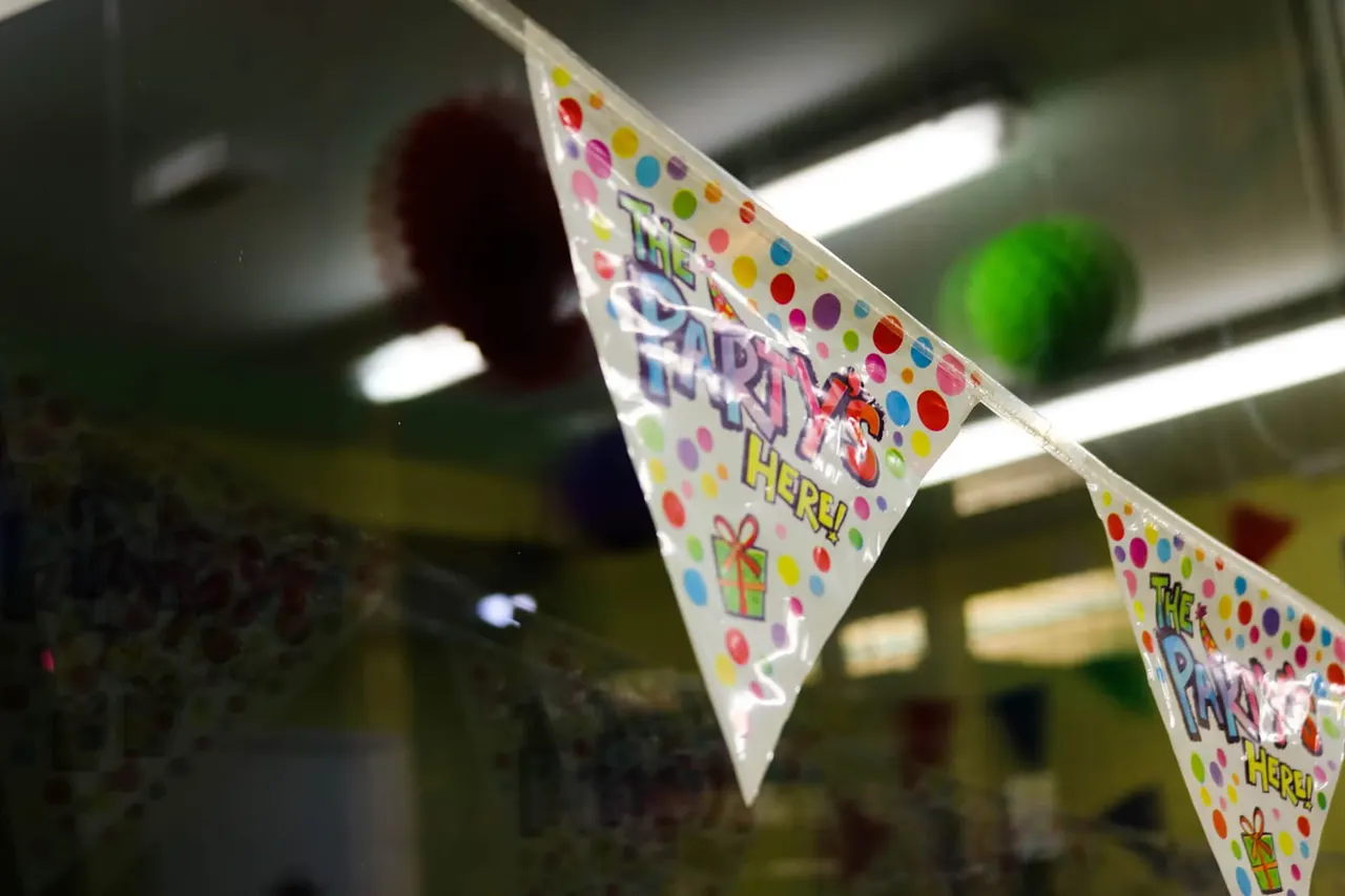 Colourful party bunting strung indoors with "The Party Here" text and multicoloured polka dots, with blurred children and play equipment behind.
