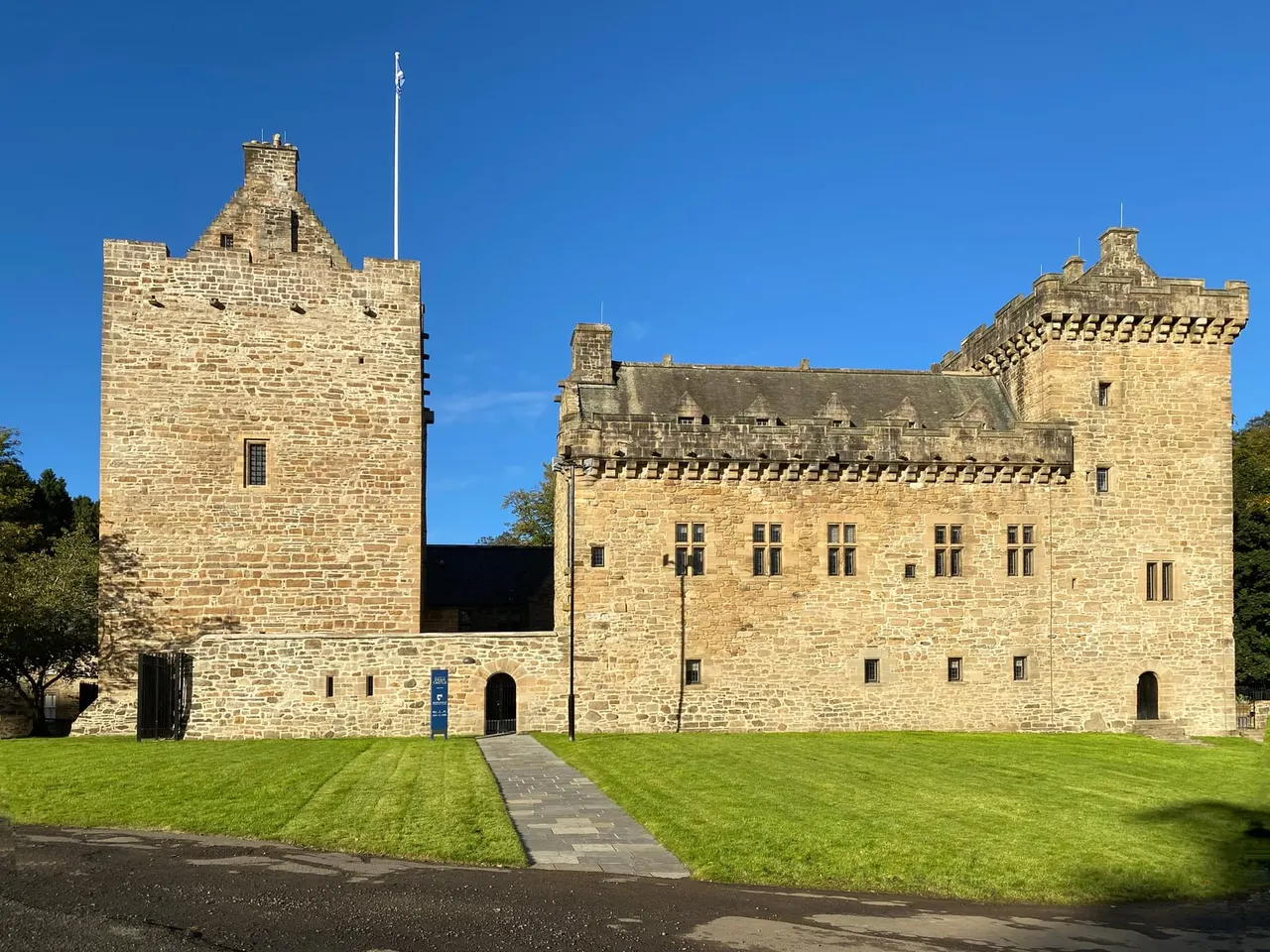 Stone castle with two towers and crenellated walls set in manicured grounds under clear blue sky.