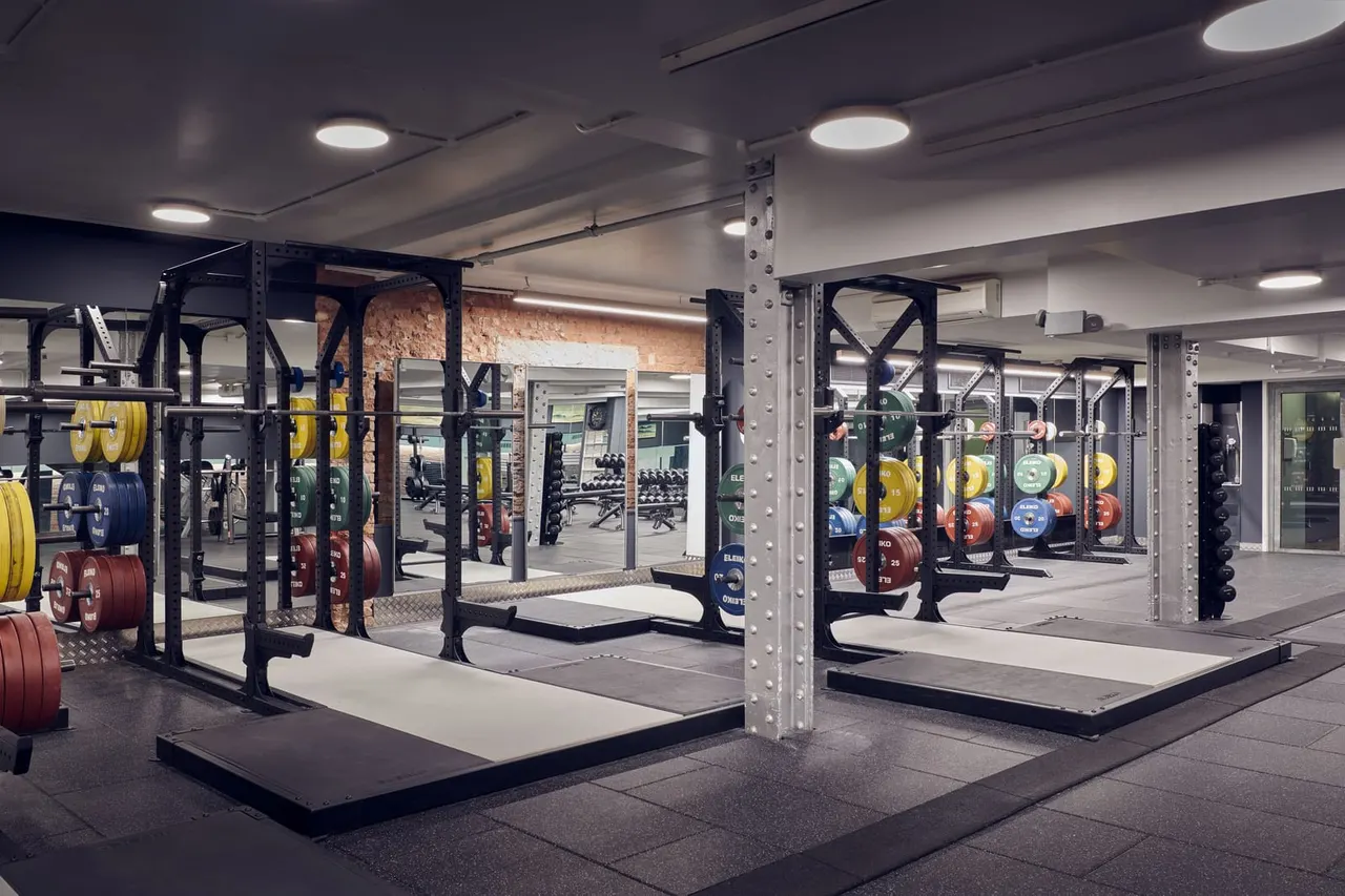Modern gym interior with weight racks, colourful plates, and lifting platforms under bright ceiling lights.