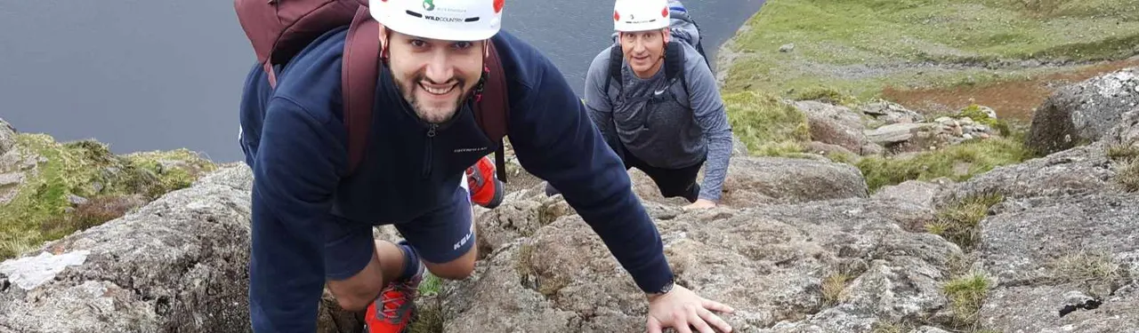 Two people in safety helmets rock climbing or scrambling up a steep rocky hillside with vegetation.