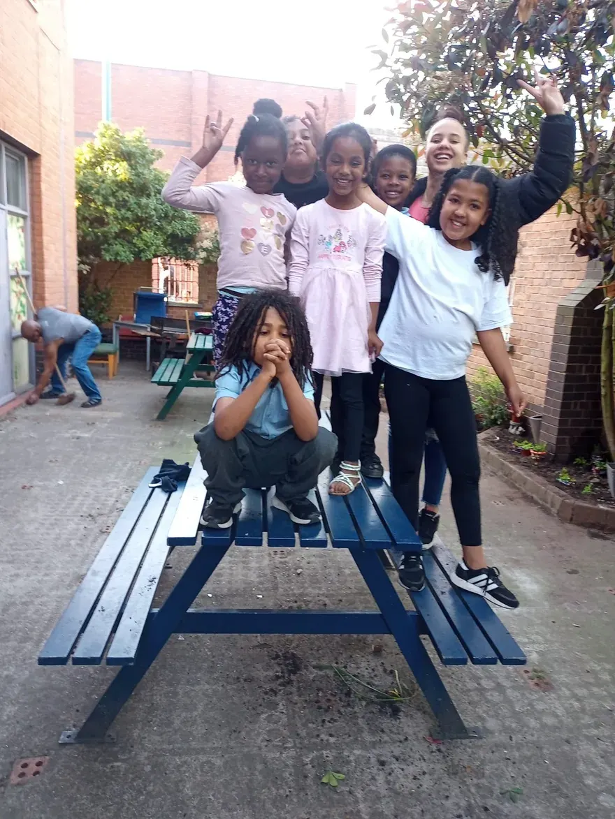 Group of children and a young adult posing together at a picnic table in a brick-walled outdoor courtyard area.